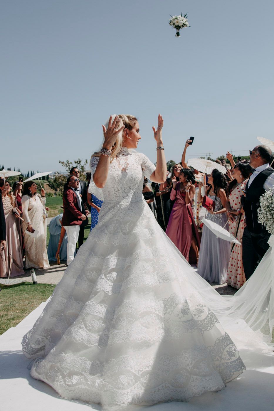 A bride in a lace wedding dress throws her bouquet high into the air as guests watch excitedly.