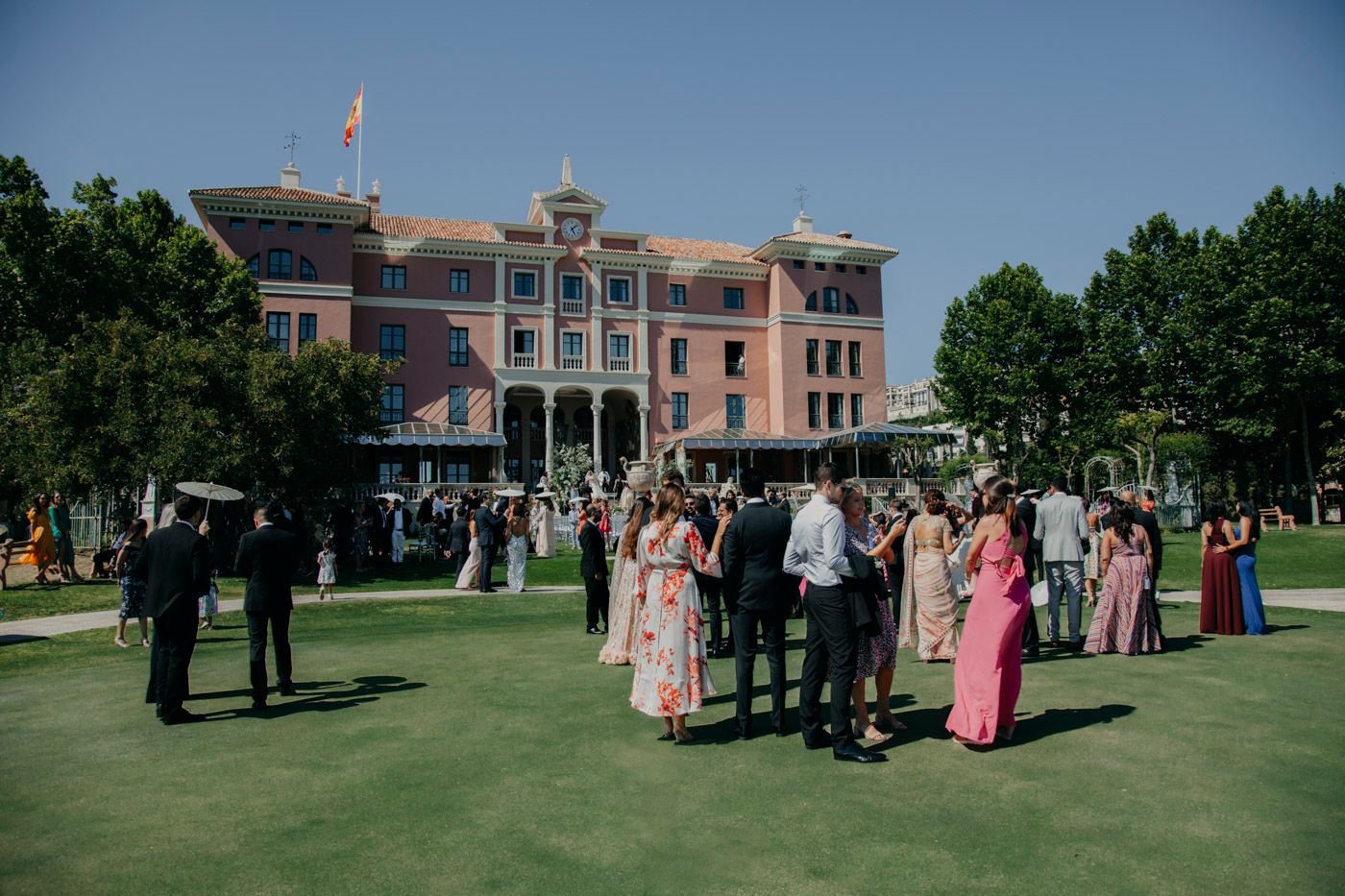 Wedding guests gather on a manicured lawn in front of a grand pink historic mansion during an elegant outdoor celebration.