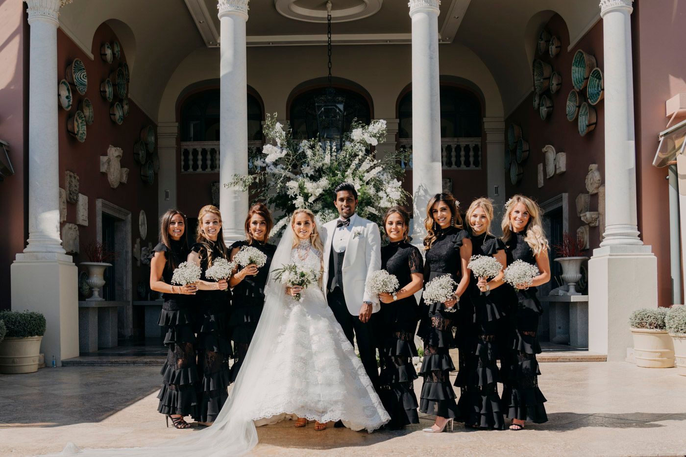 Wedding party poses in formal black and white attire on elegant colonnade steps with classical architecture backdrop.