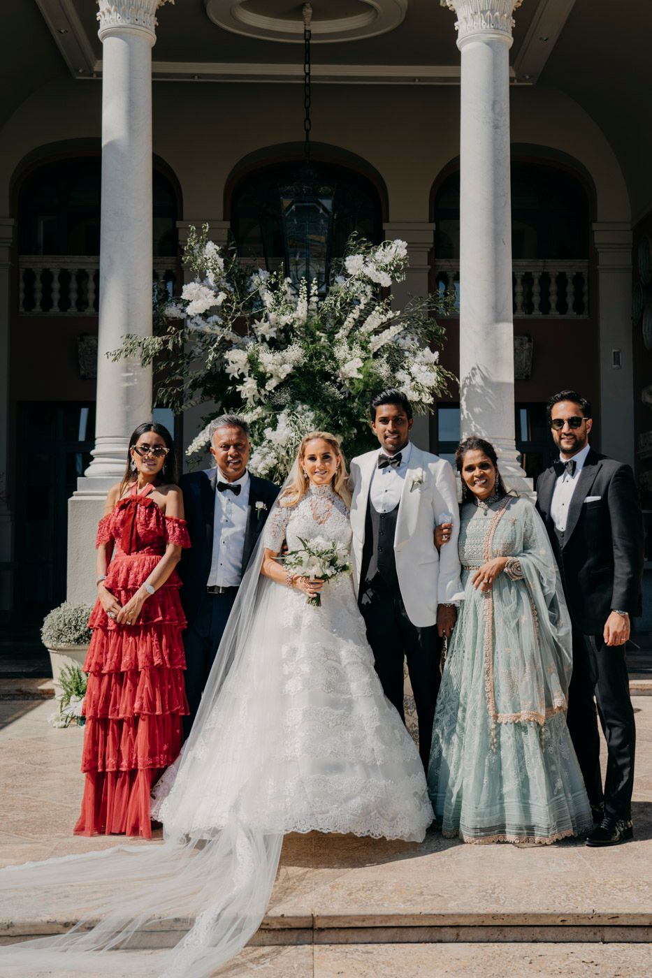 Wedding party poses at elegant venue with white columns and floral arch featuring guests in red and blue formal attire.