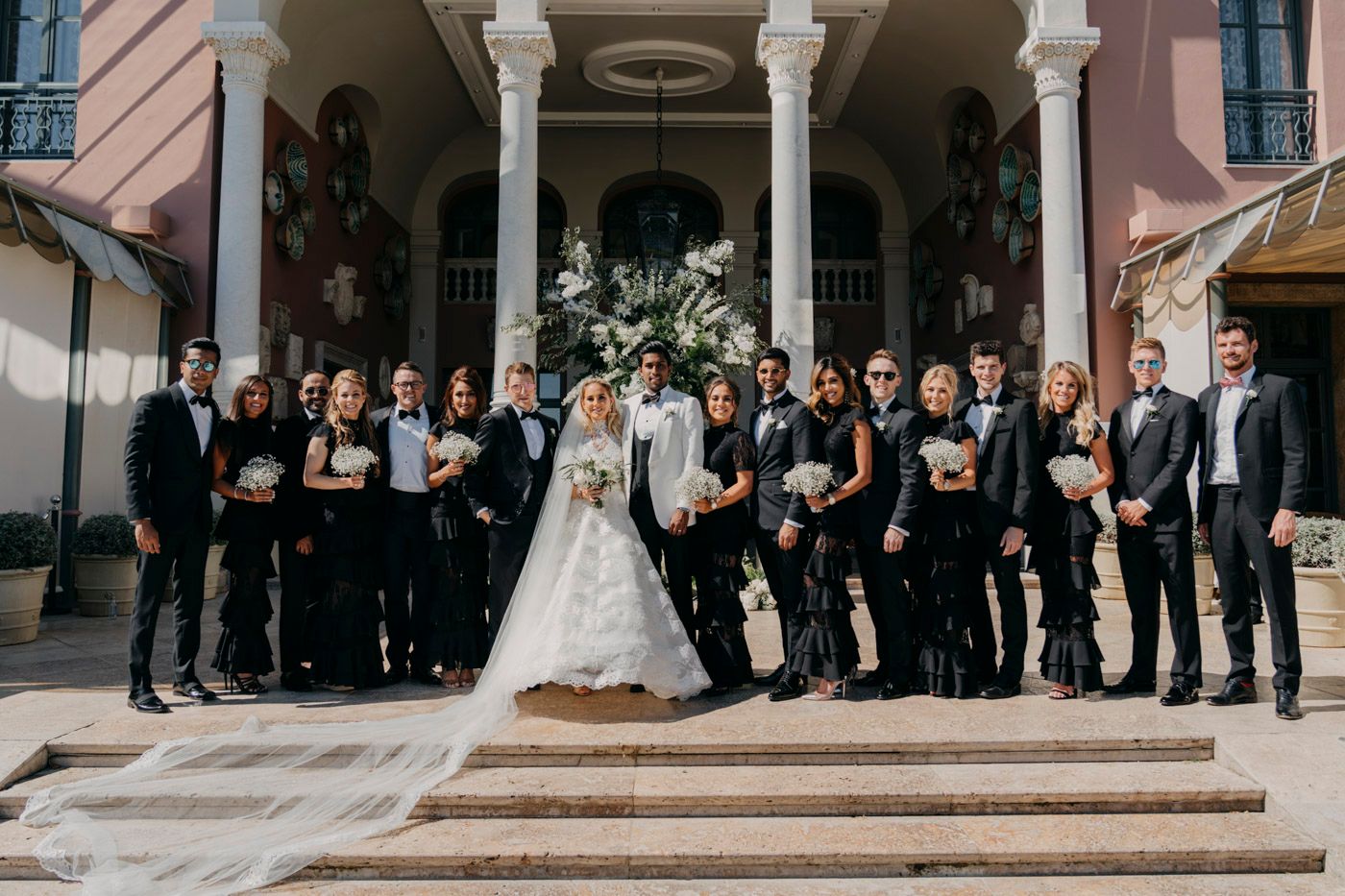 A wedding party dressed in formal black attire poses together on stone steps of a pink columned building.