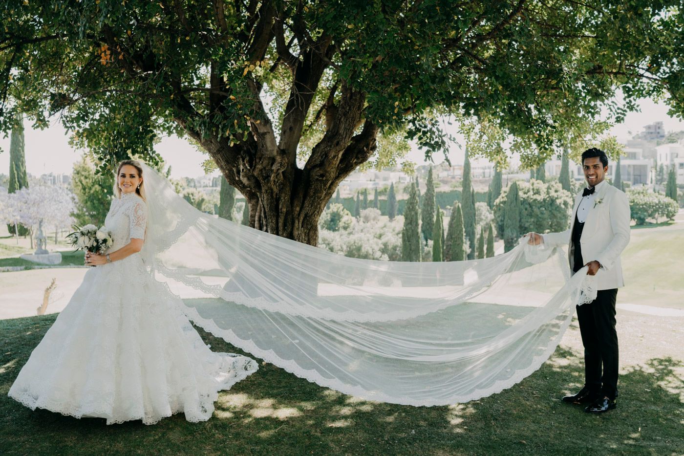 A dramatic bridal veil flows in the breeze beneath a tree during an outdoor wedding photoshoot.