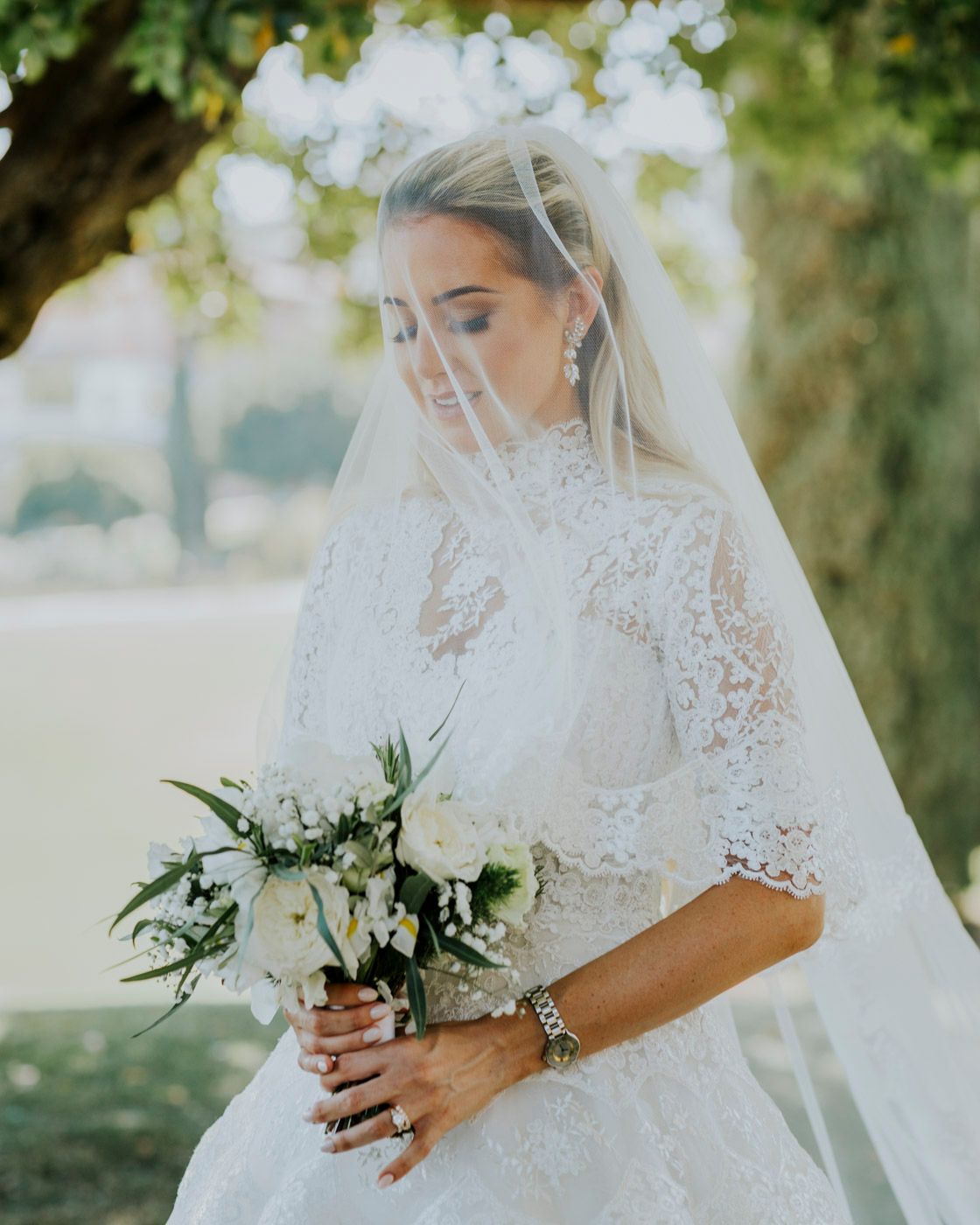 Elegant white lace wedding dress with delicate veil and classic white floral bouquet in outdoor garden setting.