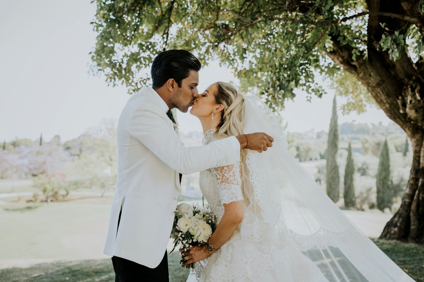 A romantic bridal couple shares a tender kiss under a blooming tree on their wedding day.