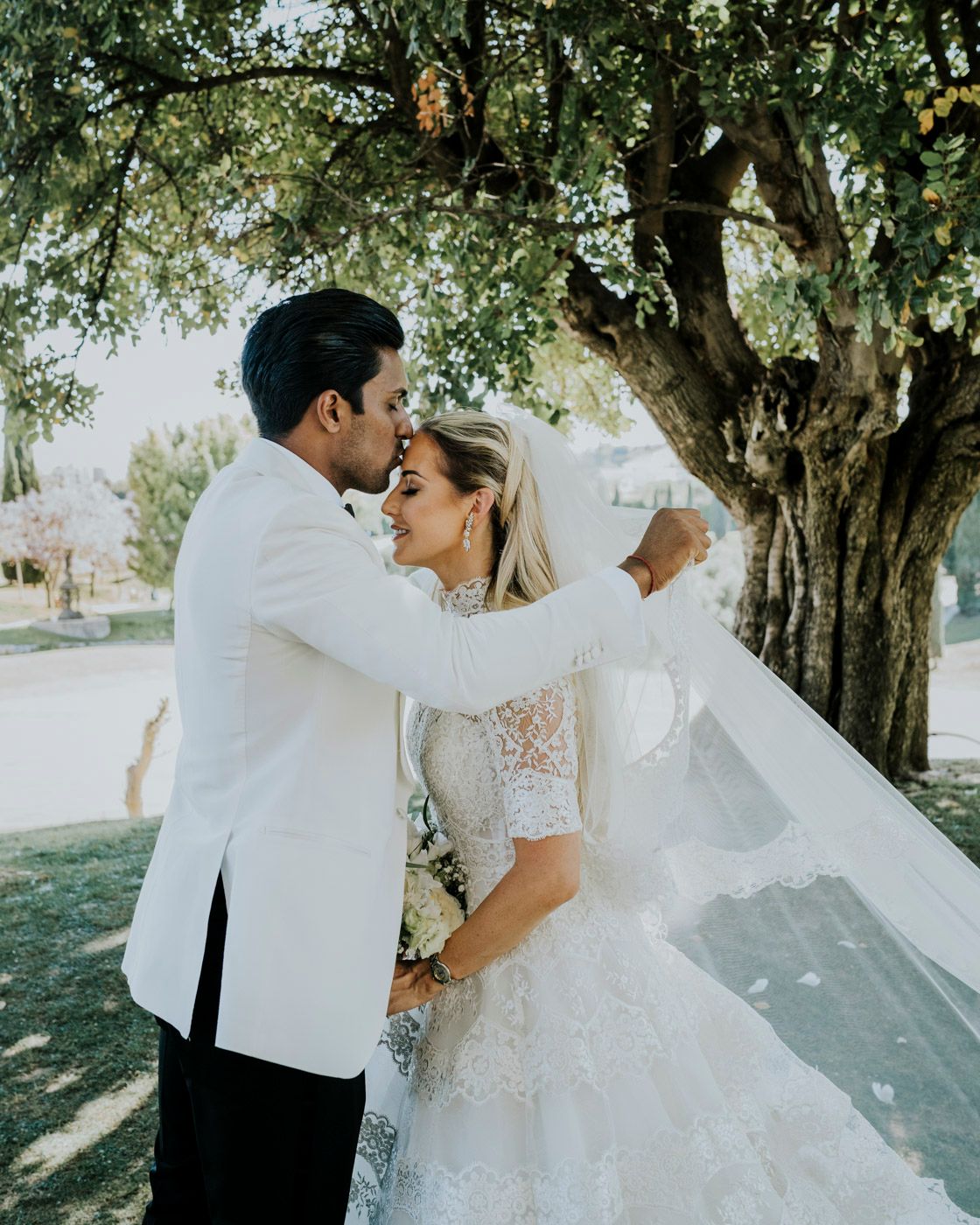 Couple embraces under a shady tree on their wedding day with veil flowing in the sunlight.