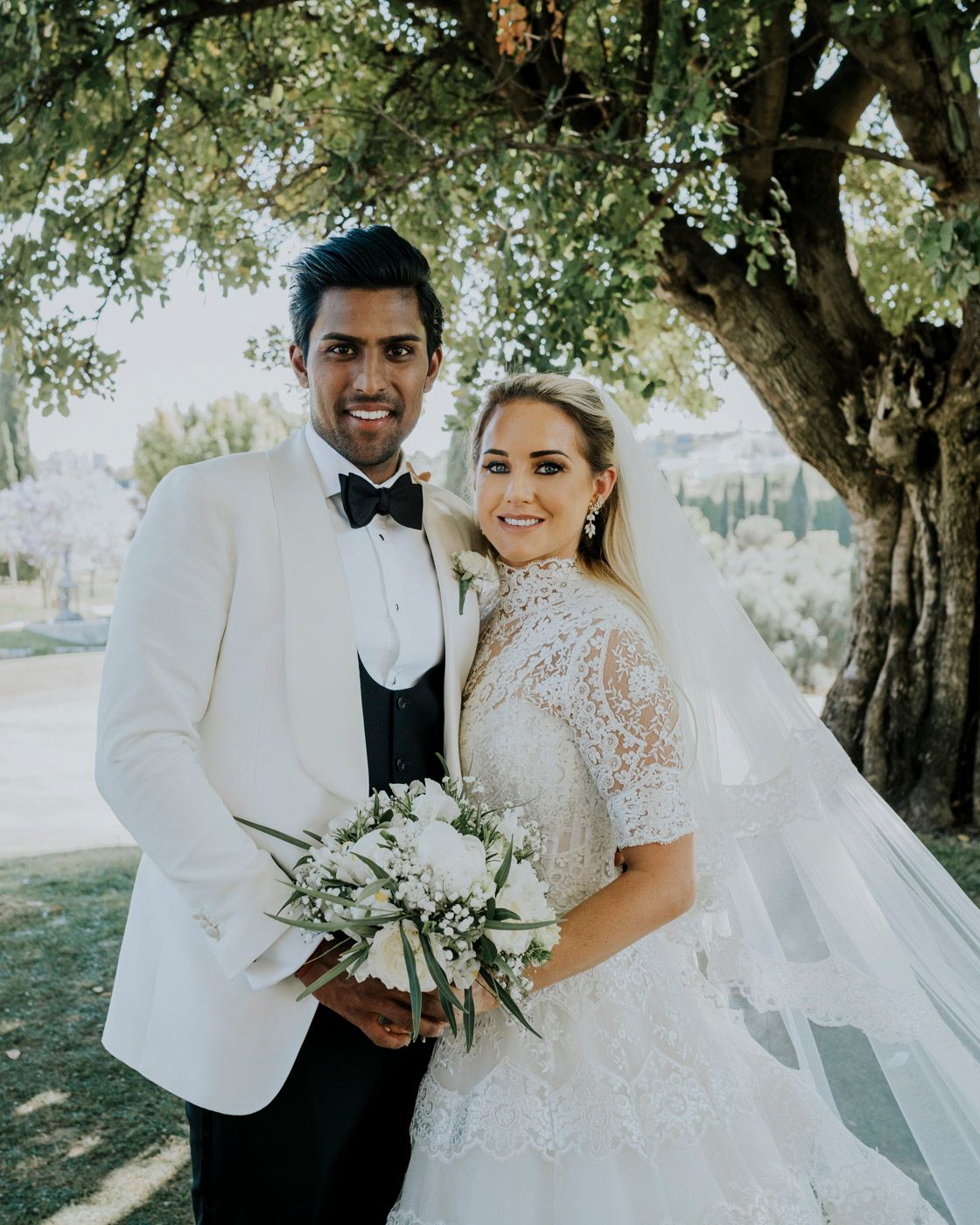 Elegant wedding portrait featuring white floral bouquet and classic ivory attire under a blossoming tree.