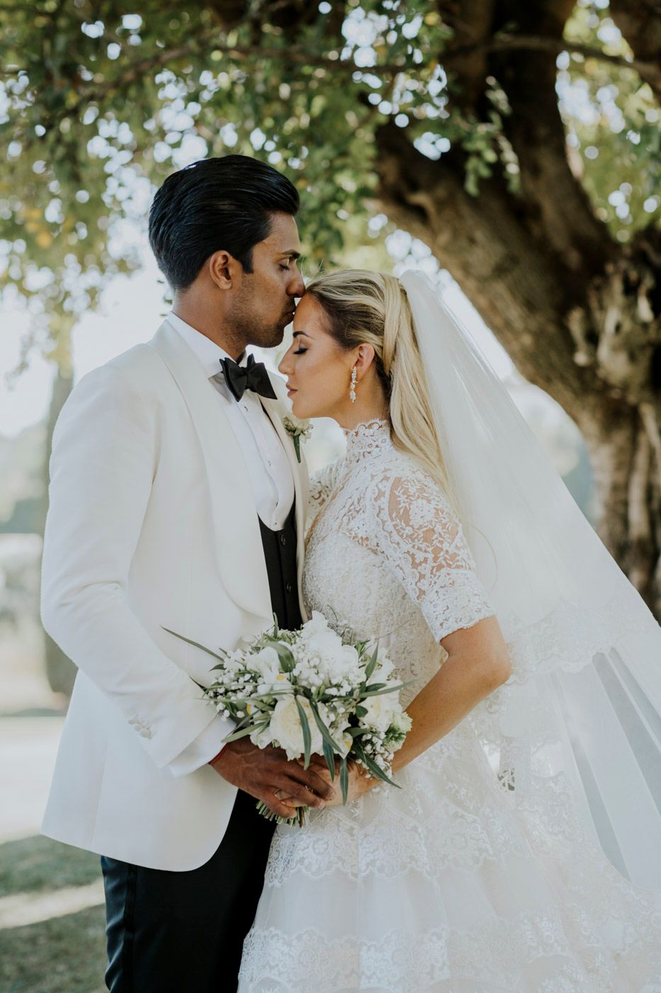 A romantic wedding couple embraces under a sunlit tree, with the bride wearing a lace gown and holding a white bouquet.