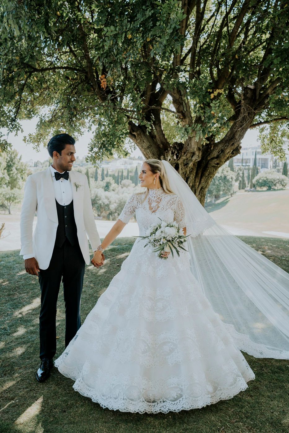 A wedding couple poses under a large tree, the bride in a lace ballgown and the groom in a white tuxedo jacket.