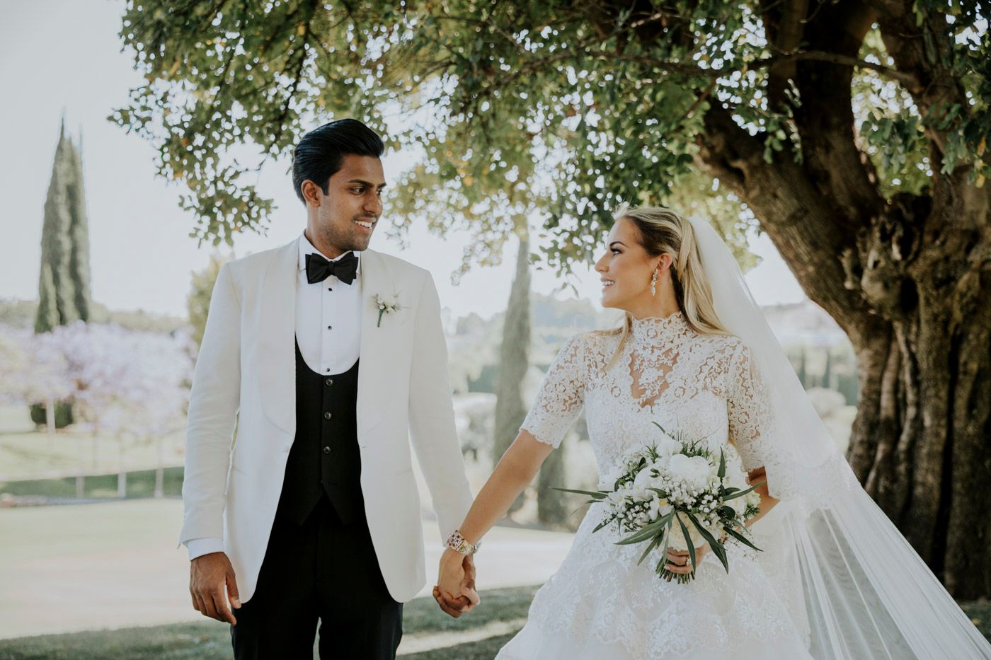 A romantic wedding couple holds hands while walking outdoors under a tree with a scenic countryside backdrop.