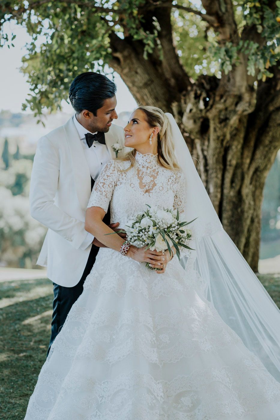 A romantic wedding couple shares an intimate moment under a large tree, with the bride wearing a lace gown and flowing veil.