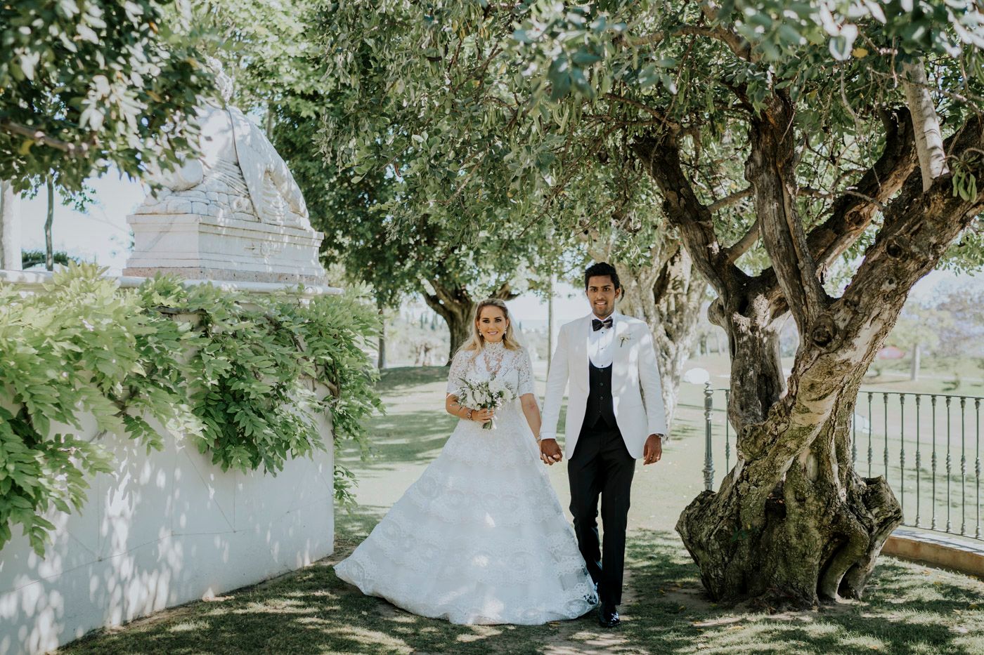A wedding couple standing together beneath an olive tree in a sunlit garden beside a white church.