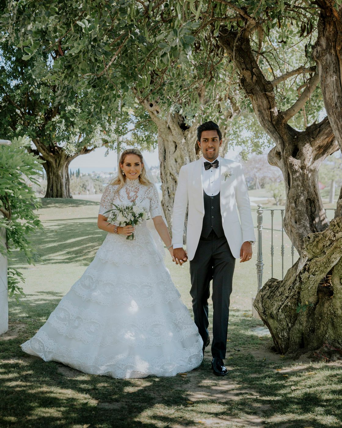 Newlyweds walk hand in hand through a garden path while the bride holds a delicate bouquet.