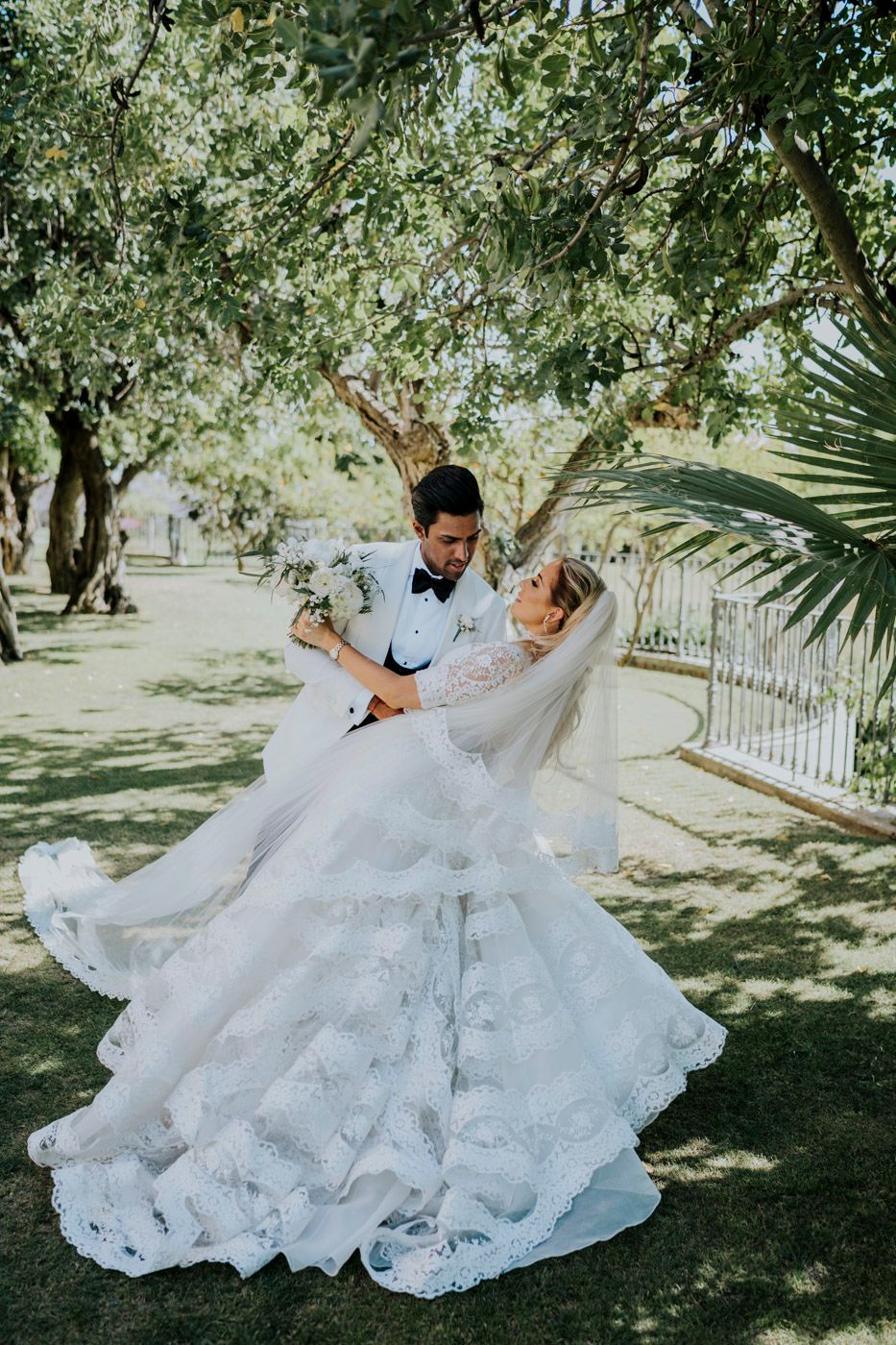 A romantic wedding couple dances together in a sunlit garden setting with lush trees and a flowing white lace gown.