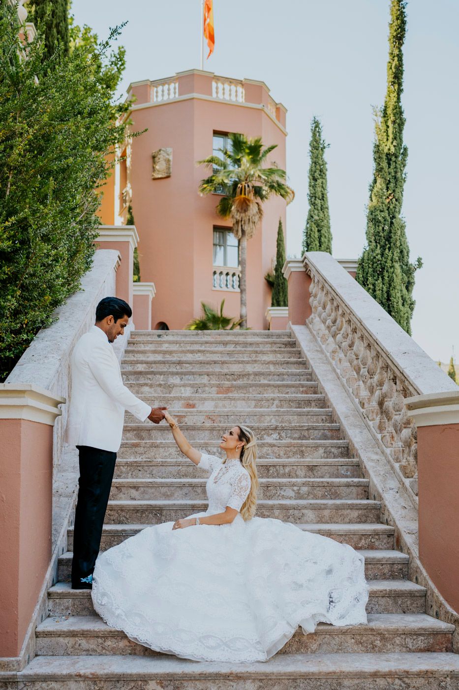A dreamy wedding scene on elegant stone stairs at a pink Mediterranean-style villa with cypress trees and palm fronds.
