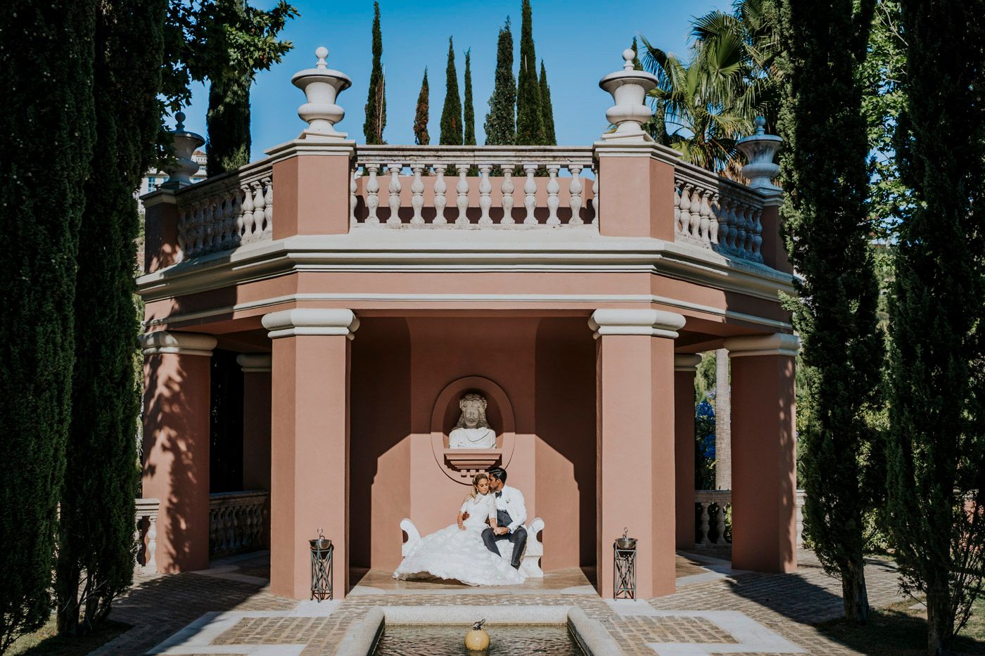 A classical pink stone gazebo with ornate architecture stands among cypress trees in a Mediterranean style garden.