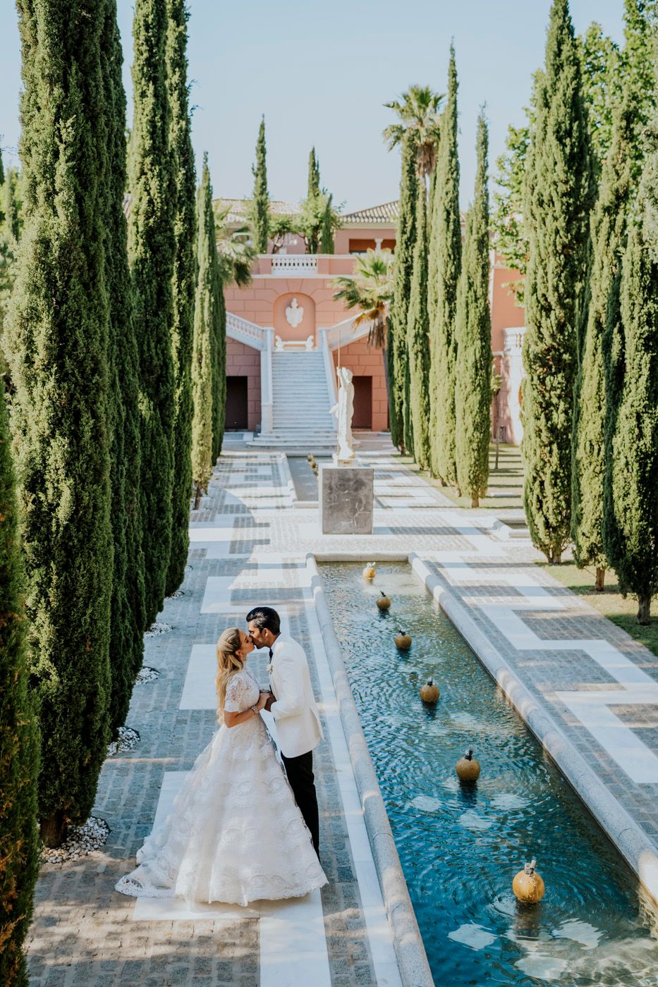 Wedding couple stands beside reflective pool lined with tall cypress trees at elegant Mediterranean-style venue.