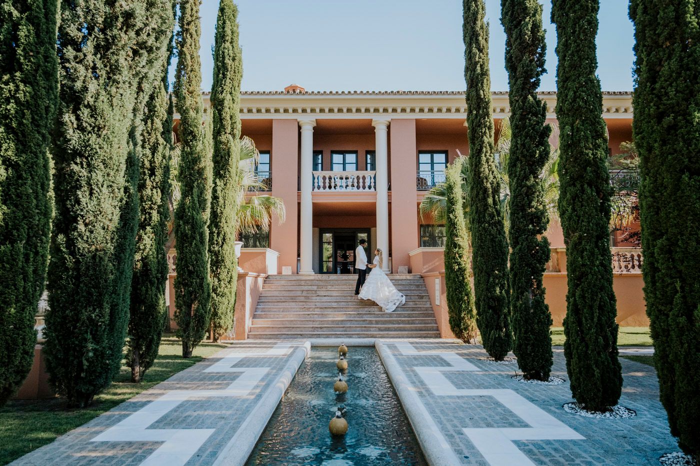 Mediterranean villa entrance with cypress trees lining a water feature leading to pink facade and grand staircase.