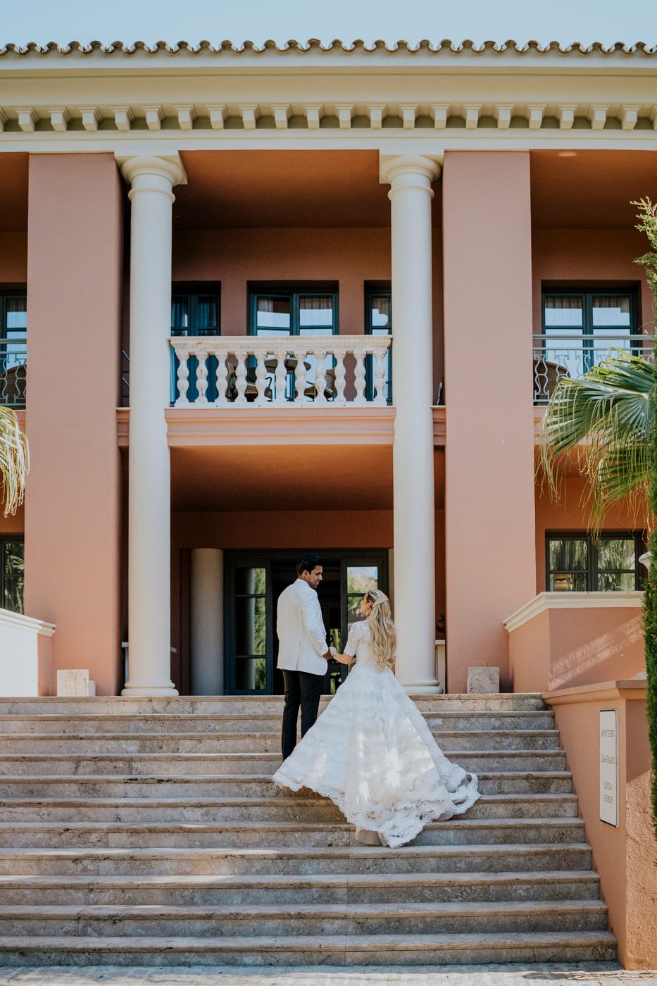 An elegant wedding couple stands at a pink Mediterranean villa with white columns and ornate stone balustrades.
