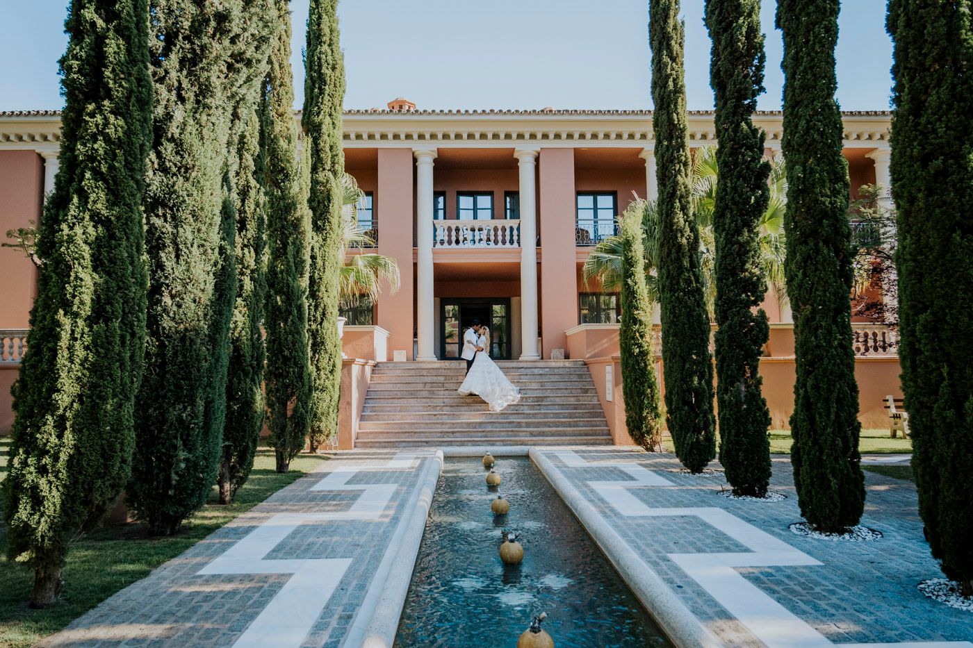 Elegant Mediterranean villa with cypress trees lining a reflecting pool leading to grand entrance steps.
