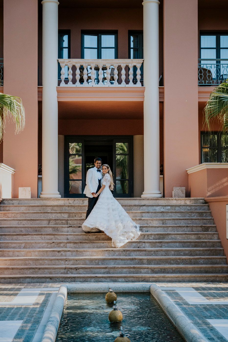 Elegant couple stands on grand marble staircase with white columns and fountain at pink Mediterranean-style villa.