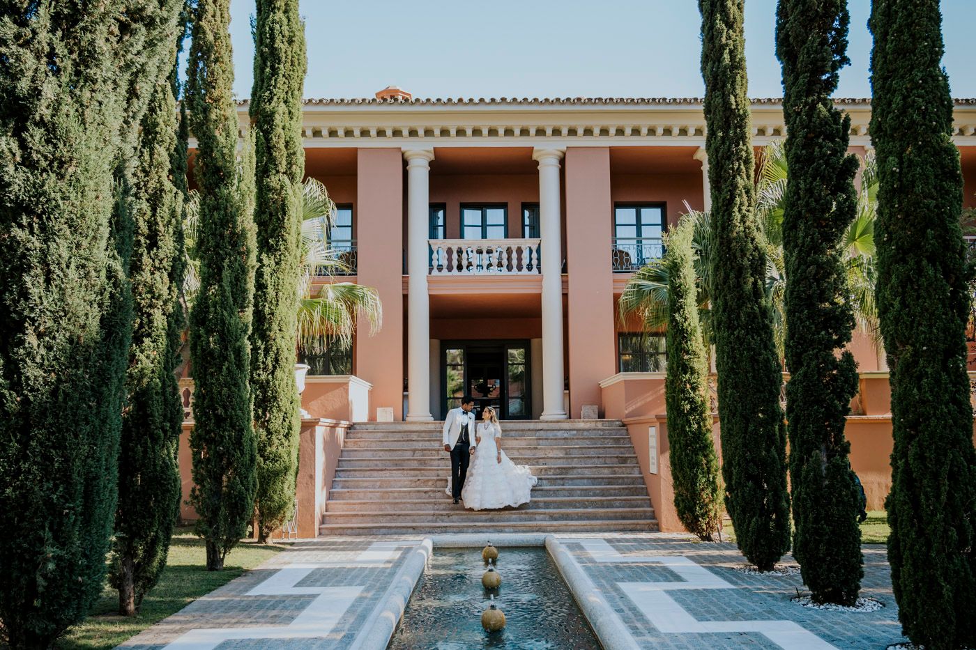 A Mediterranean-style villa with terracotta walls and tall cypress trees frames a reflective pool leading to grand entrance steps.