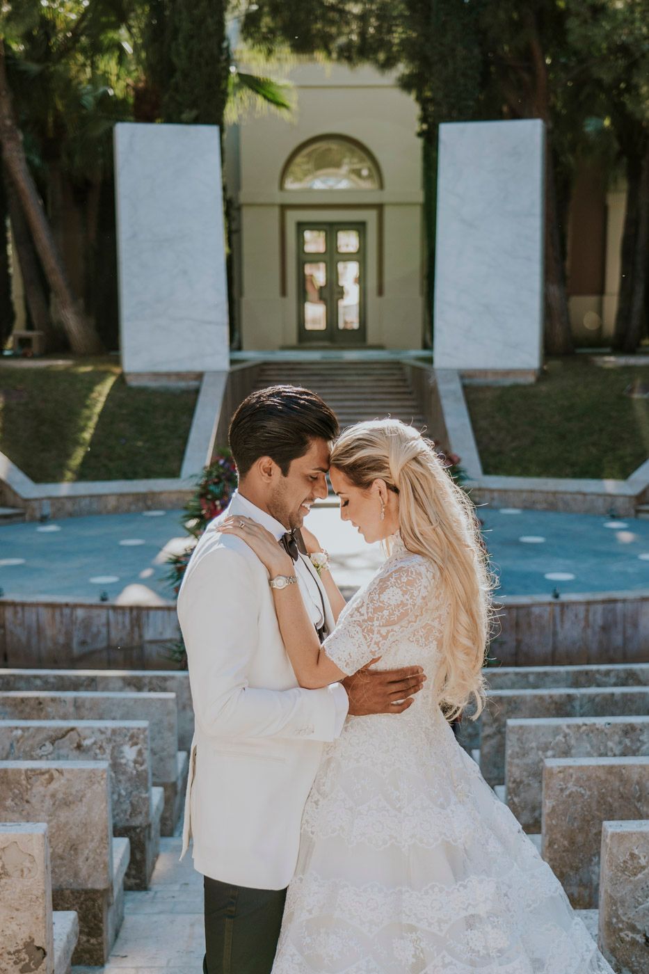 A romantic wedding couple embraces on stone steps in front of an elegant arched doorway entrance.