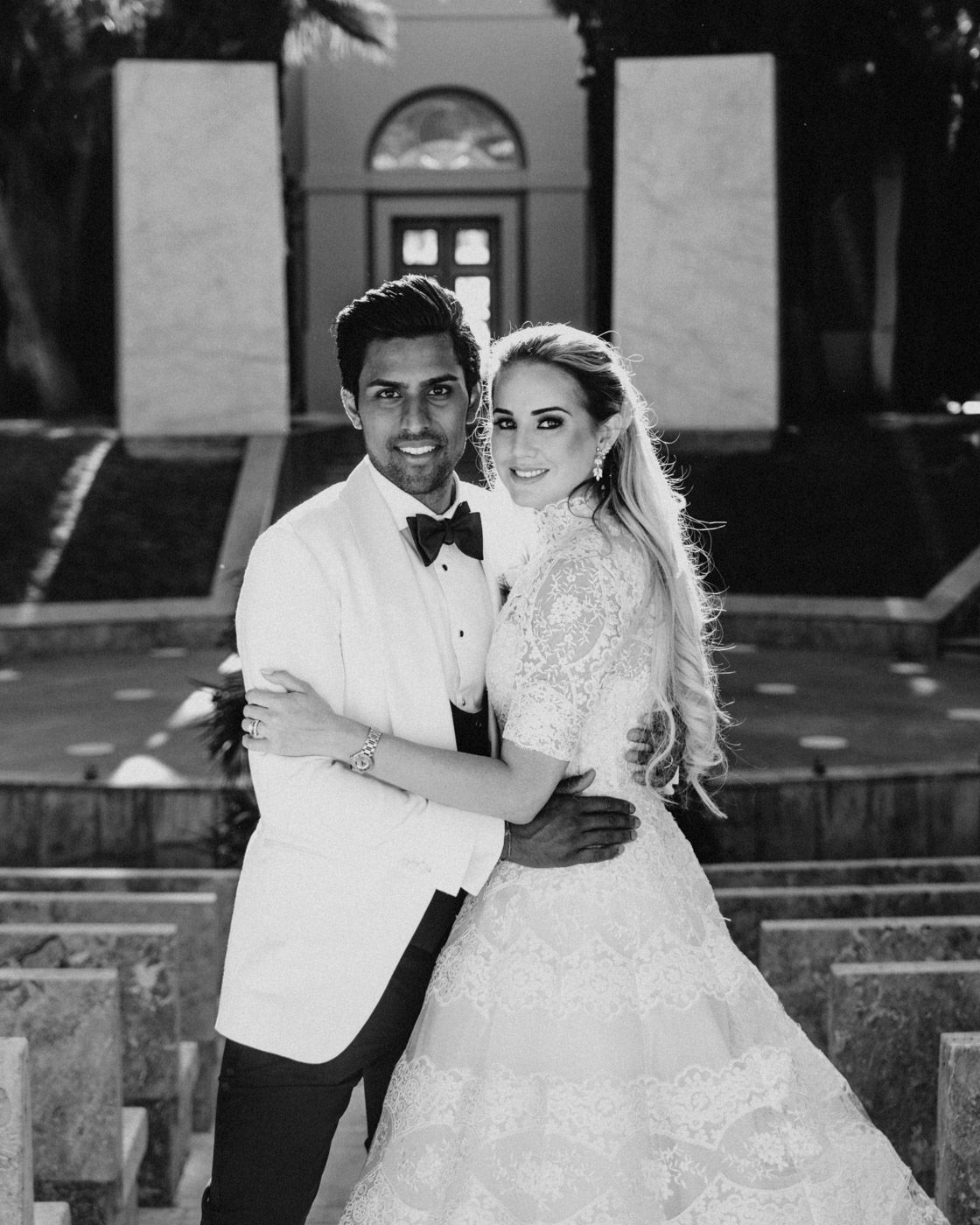 A black and white wedding portrait of a couple in formal attire standing in front of a grand building entrance.