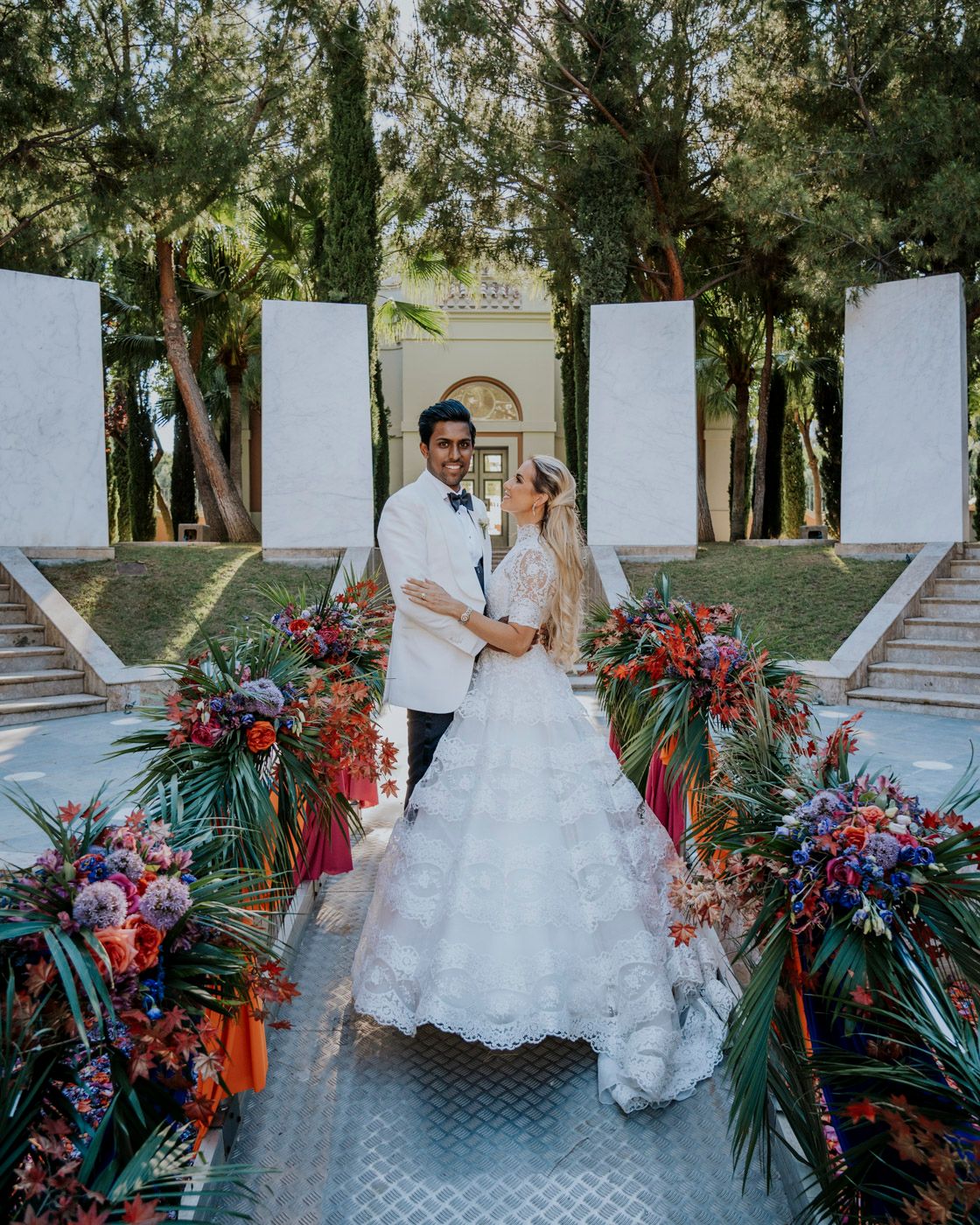 A couple stands on a tropical-decorated walkway with lush florals and palm arrangements for an outdoor wedding ceremony.