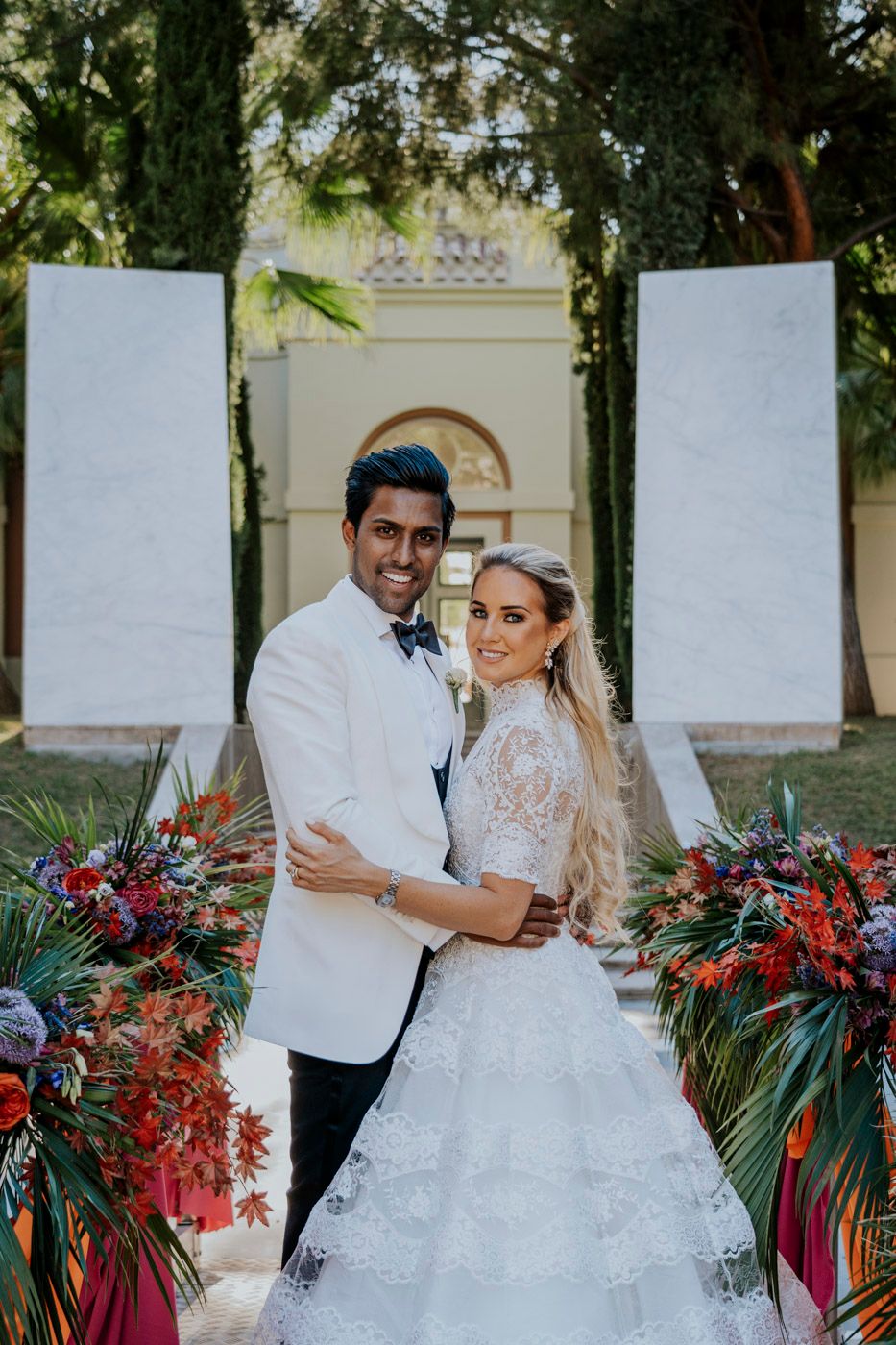 A romantic outdoor wedding scene with vibrant red floral arrangements and white decor elements near a grand archway.
