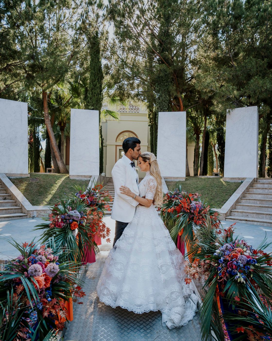 Couple shares an intimate moment on outdoor wedding steps decorated with vibrant tropical florals and greenery.