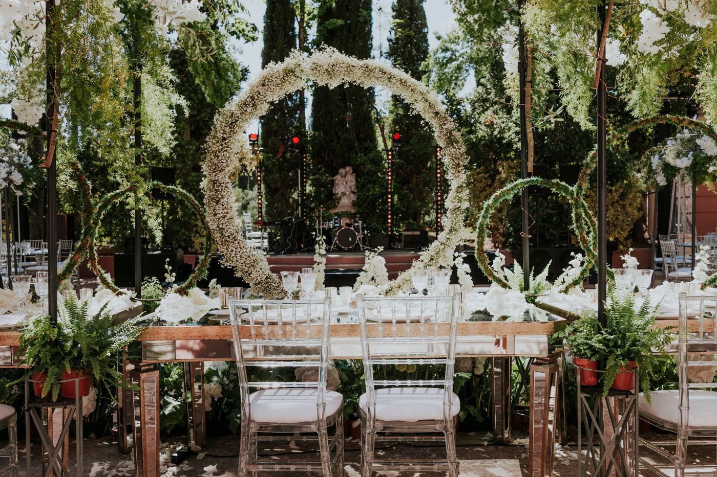 Elegant outdoor wedding reception setup with white floral arches and garden chairs in a wooded setting.