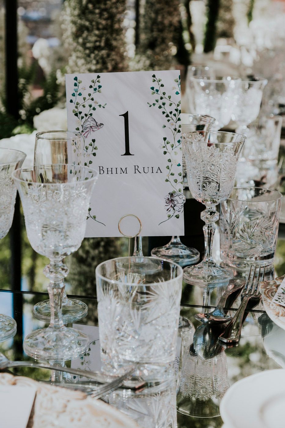 Elegant crystal glassware surrounds a delicate floral table number card displaying '1 Bhim Ruia' at a wedding reception.
