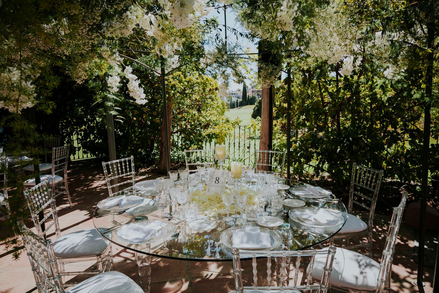 Elegant outdoor wedding reception tables set with glassware under natural green foliage and string lights.