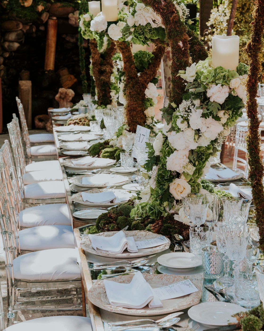 Rustic outdoor wedding reception table with white floral arrangements, crystal glassware and natural wooden chairs.