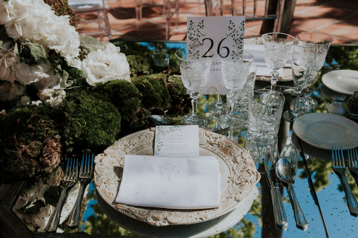 A rustic wedding table setting with moss decor, white roses, and elegant table number twenty-six.