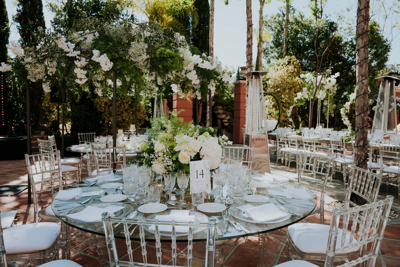 Elegant wedding reception table setting with white floral centerpieces and silver chiavari chairs in outdoor venue.