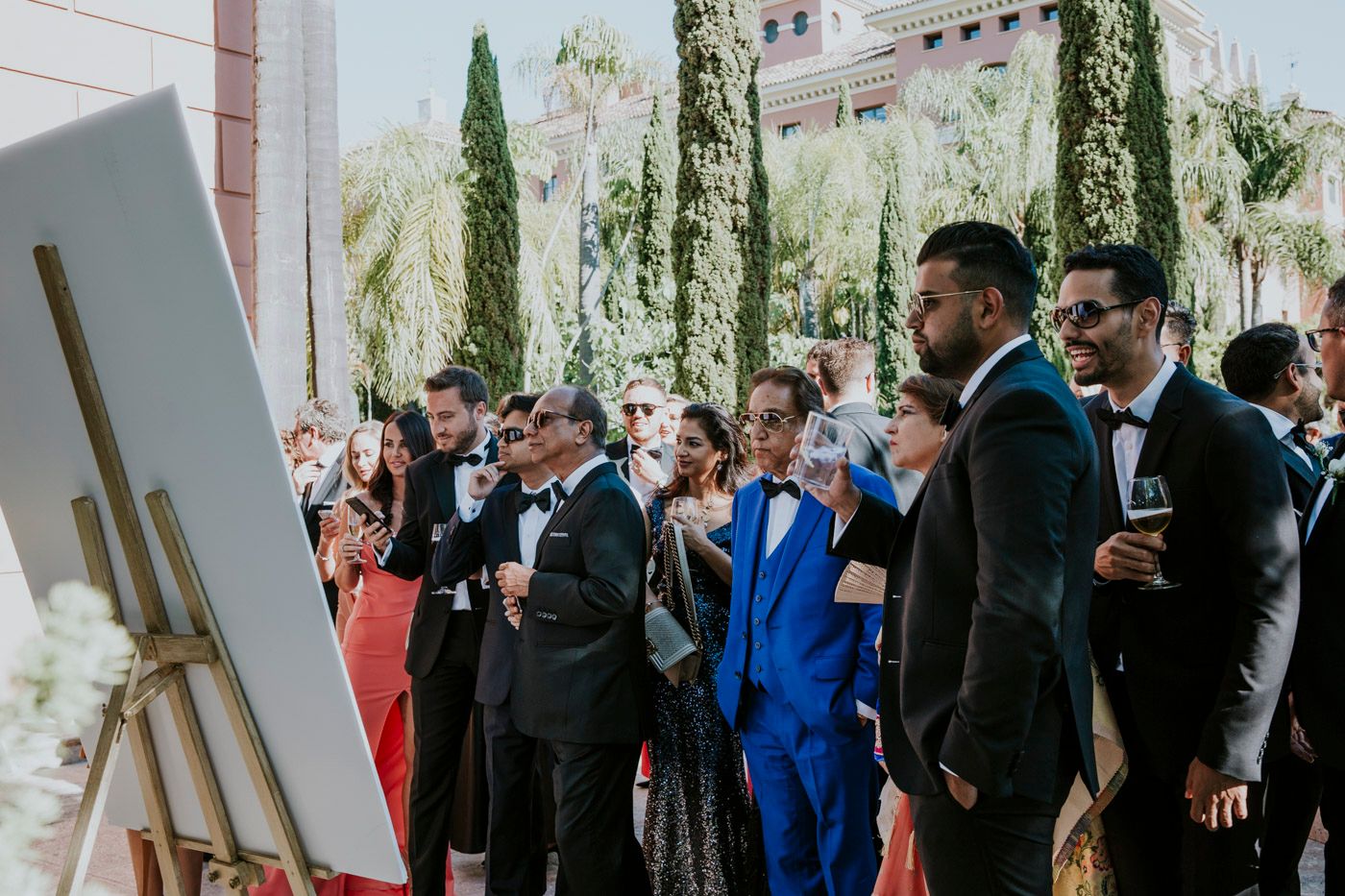 Formal event attendees in suits gather around an easel display at an outdoor venue with cypress trees.