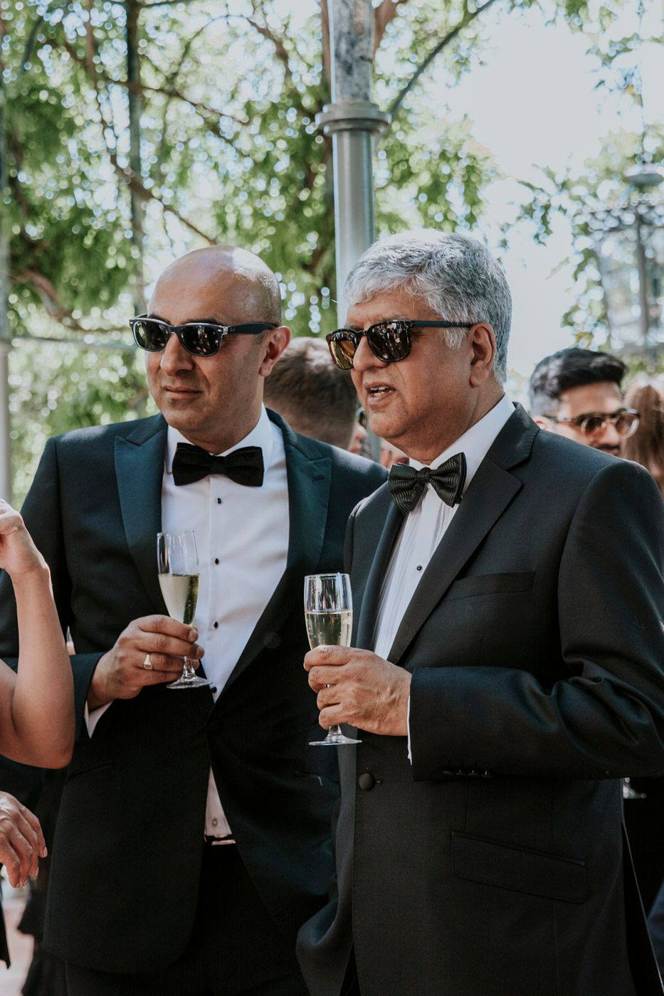 Two guests in formal black tuxedos and bow ties hold champagne glasses during an elegant outdoor celebration.
