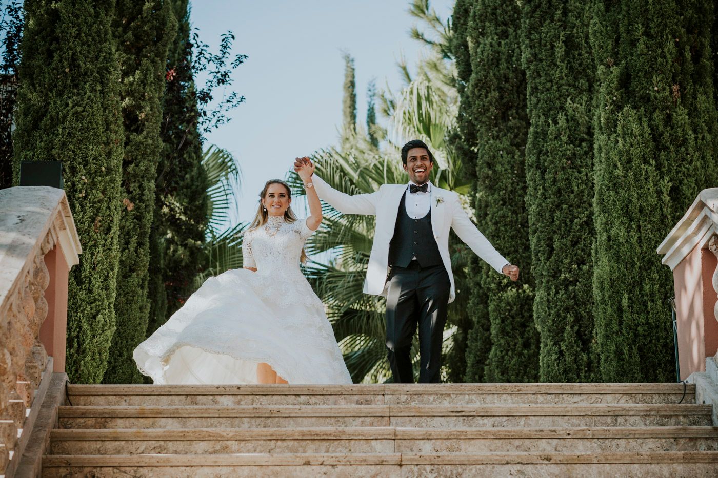 Joyful couple jumping on stone steps with tall cypress trees in their wedding attire at a garden venue.