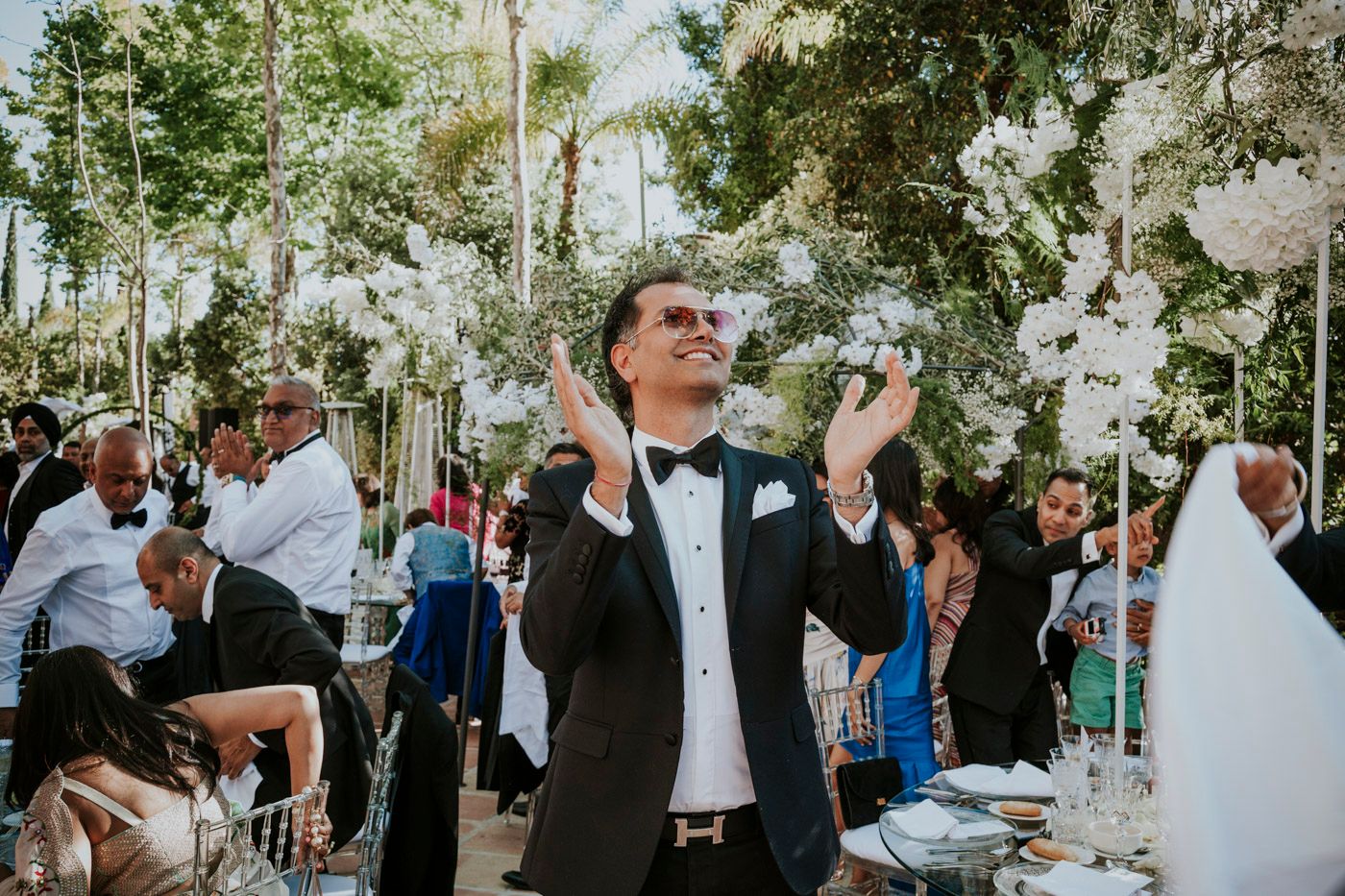 Wedding guest in a black tuxedo stands among white floral decorations at an outdoor garden reception.