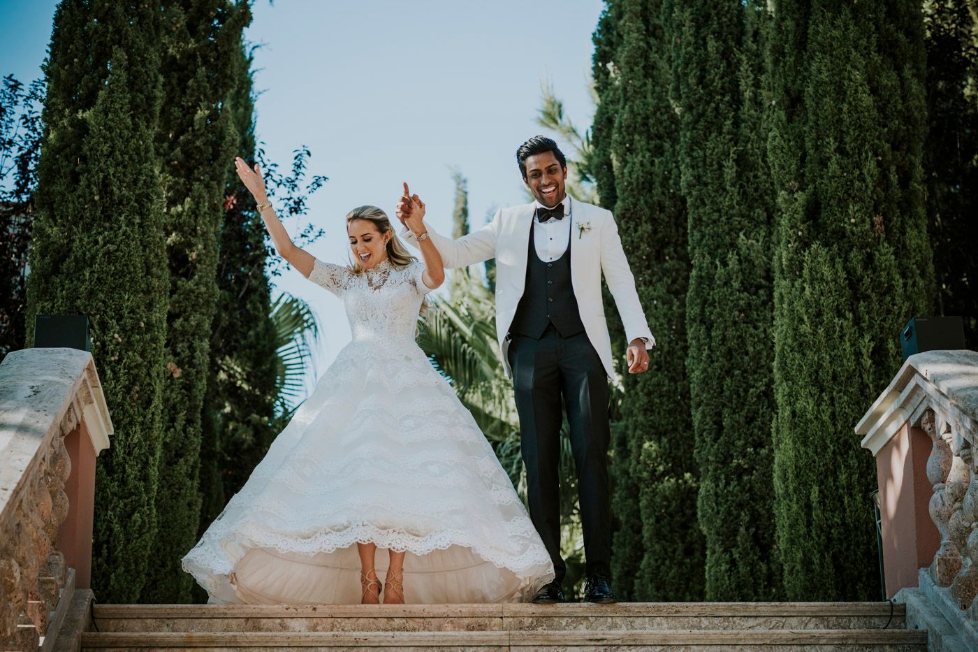 A newlywed couple celebrates joyfully at the top of ornate stone steps framed by tall cypress trees.