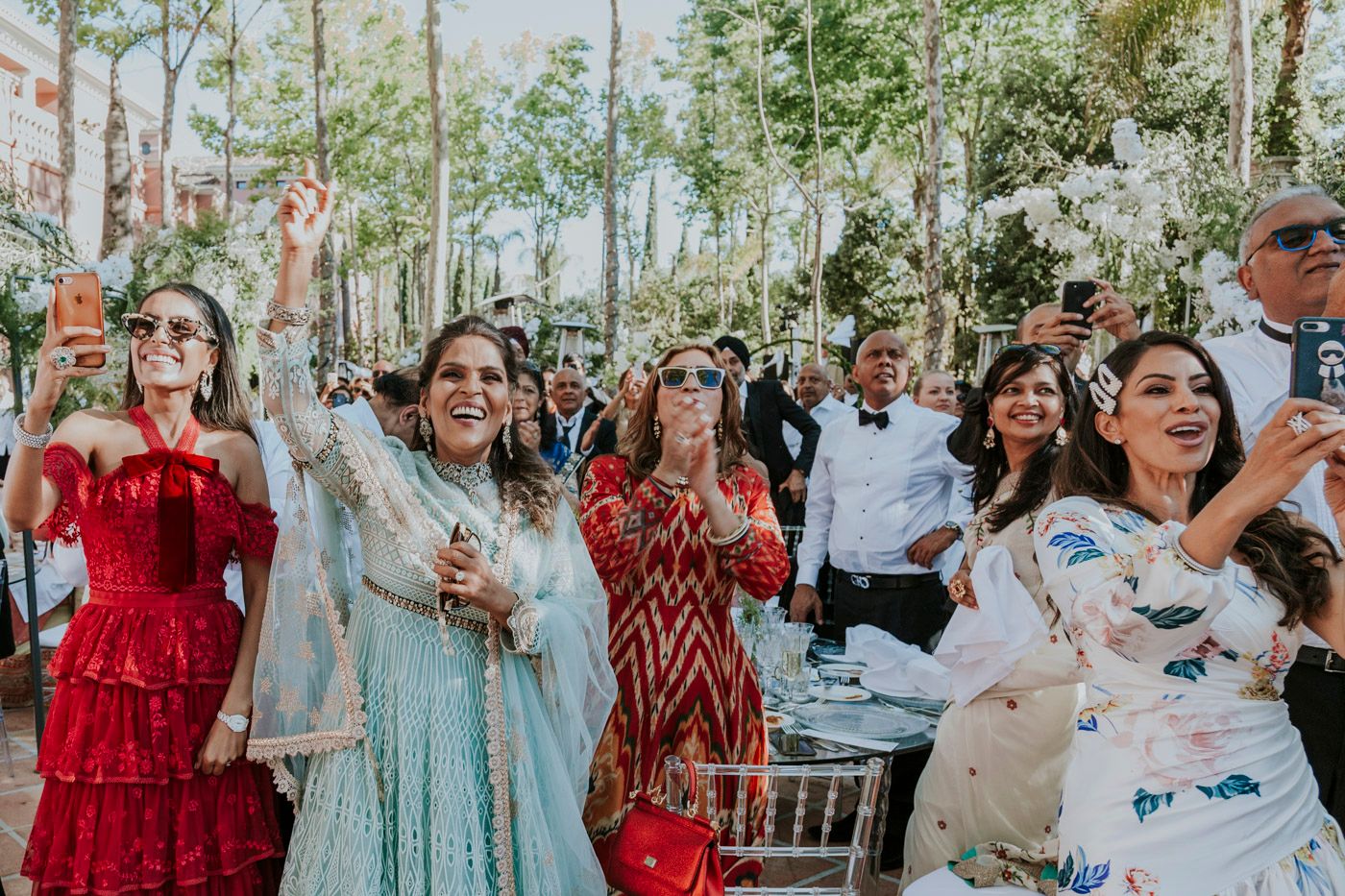 Wedding guests in colorful dresses celebrate joyfully during an outdoor reception under trees.