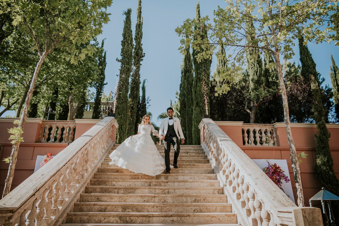 A bride and groom descend grand stone steps surrounded by towering cypress trees in a romantic garden setting.