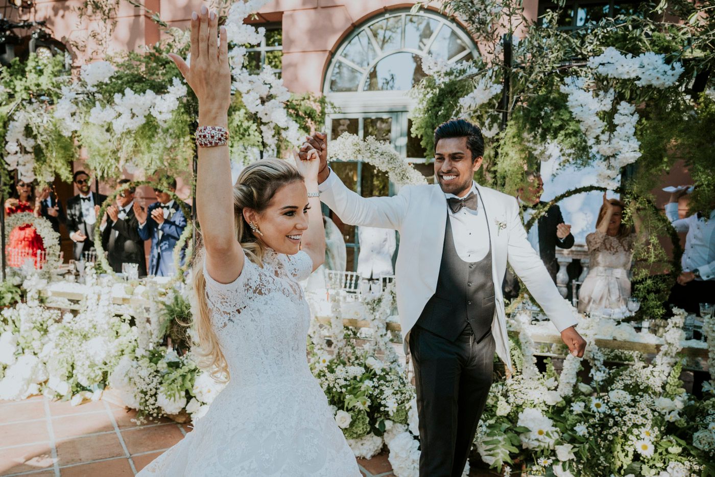 A joyful newlywed couple celebrates with raised hands during their garden wedding ceremony surrounded by white flowers.