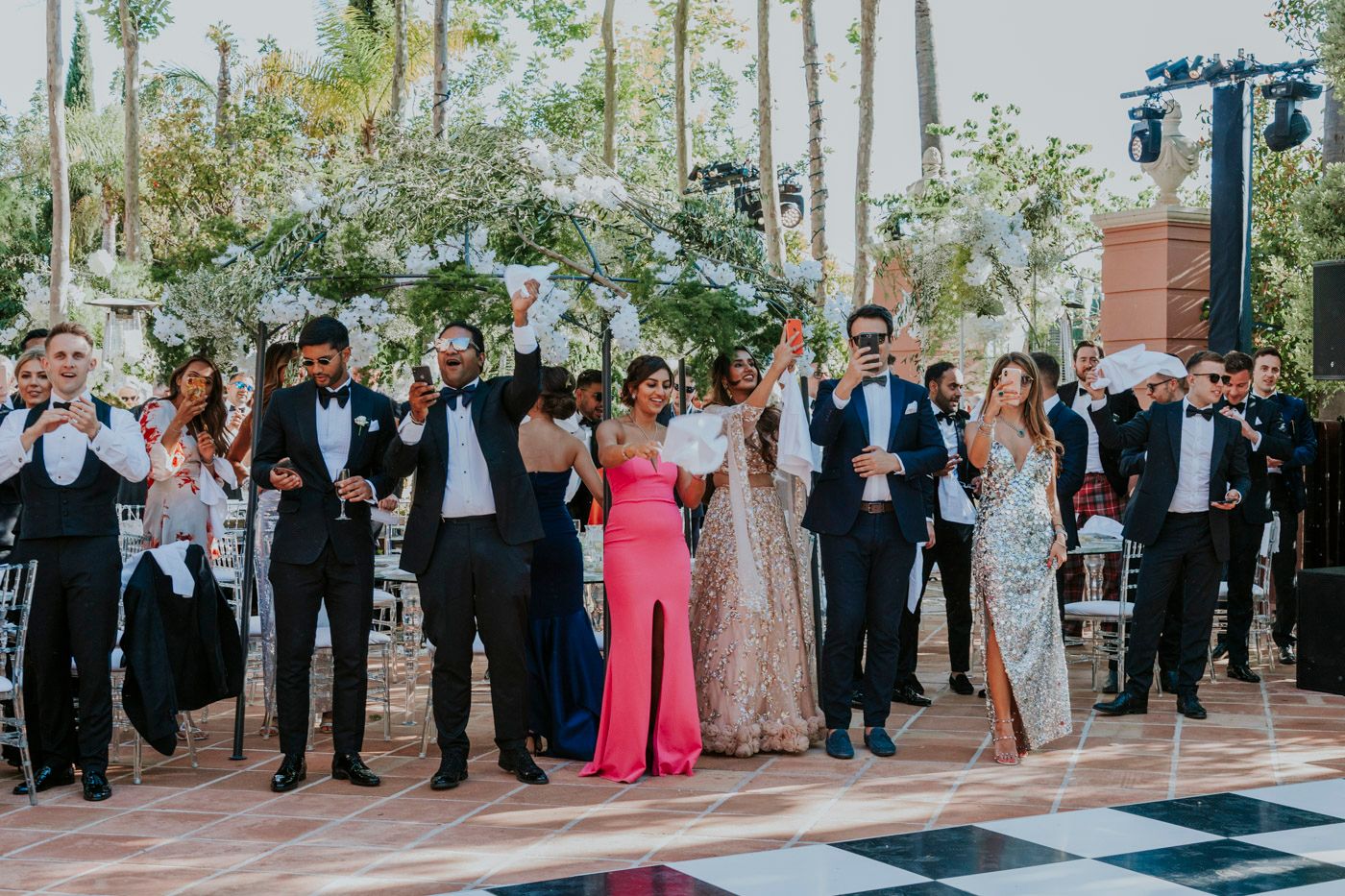 Wedding guests in formal attire raise their glasses in celebration on a black and white checkered dance floor at an outdoor venue.