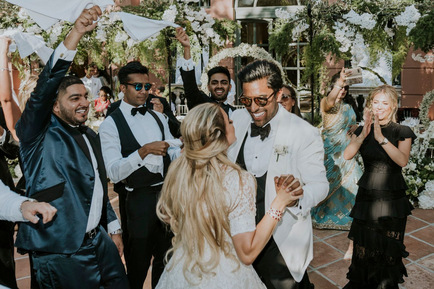 A joyful wedding reception moment with guests celebrating and dancing together under greenery decorations.
