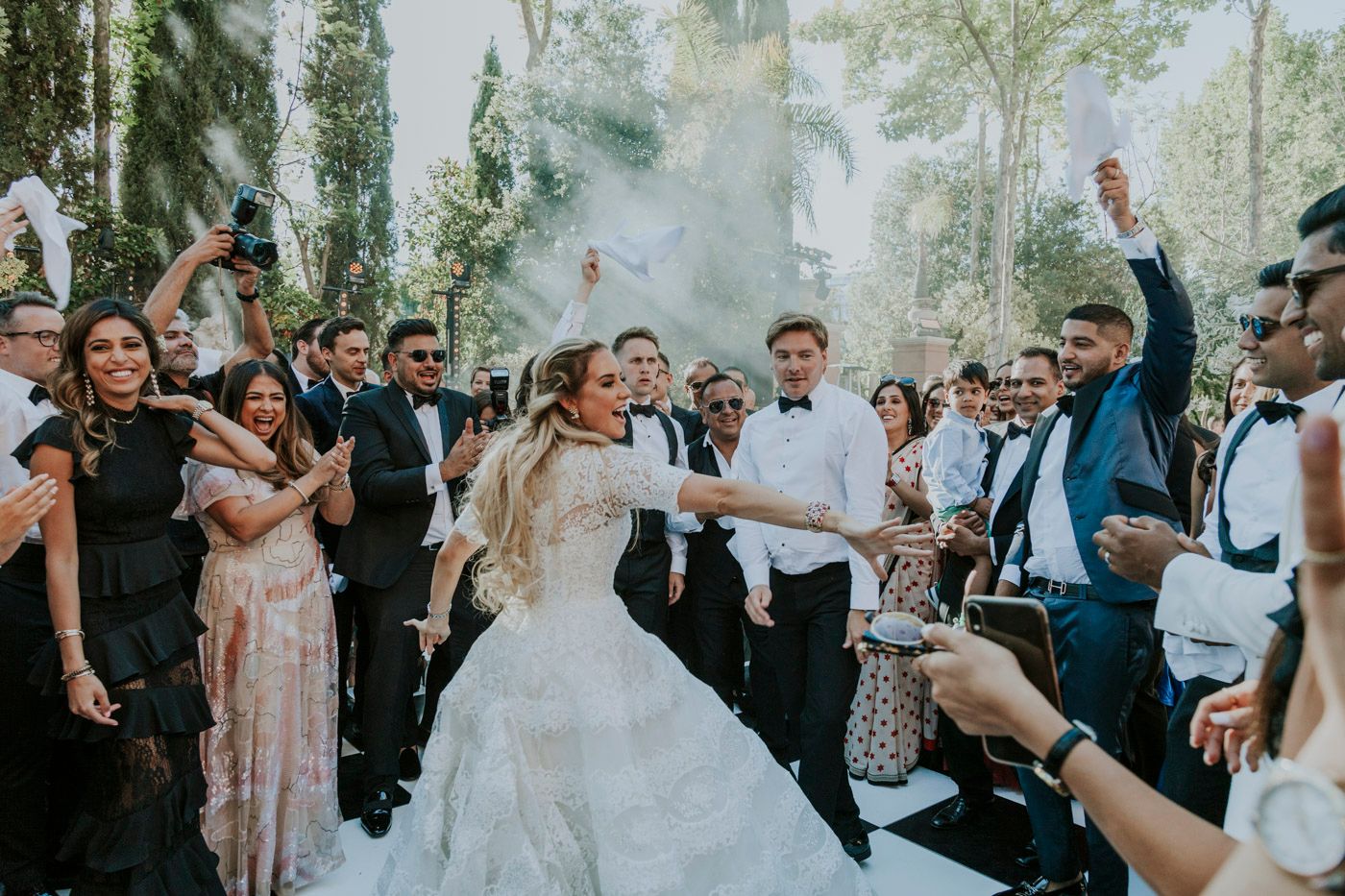Wedding party celebrates outdoors with smoke effects and joyful dancing in a forest setting during reception festivities.