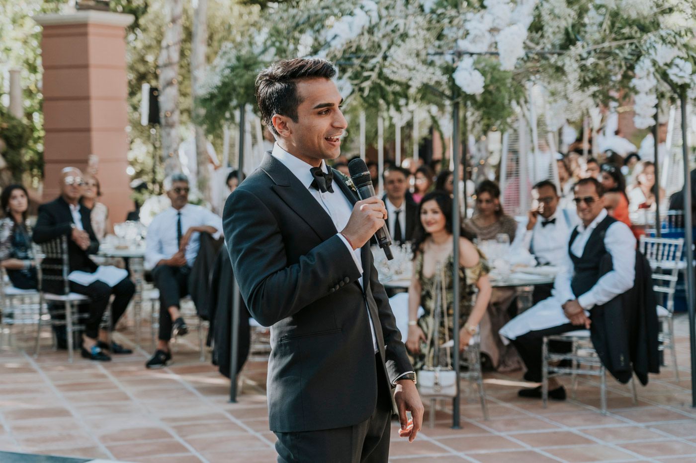 A person in a black tuxedo gives a speech with a microphone at an outdoor event while guests listen in the background.