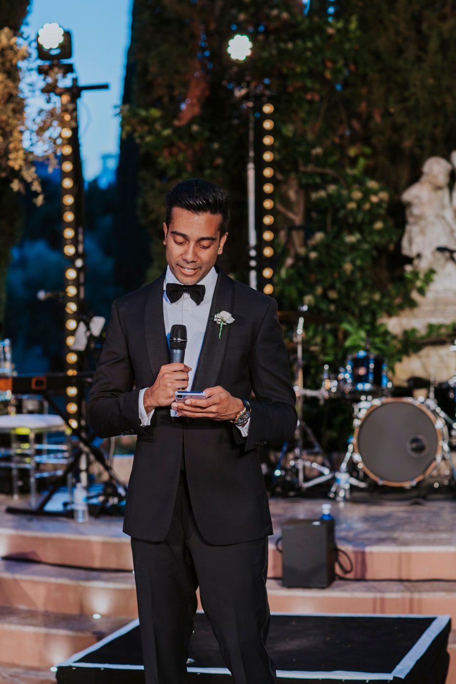 A person in a black tuxedo and bow tie delivers a speech at an outdoor wedding reception with a band setup visible.