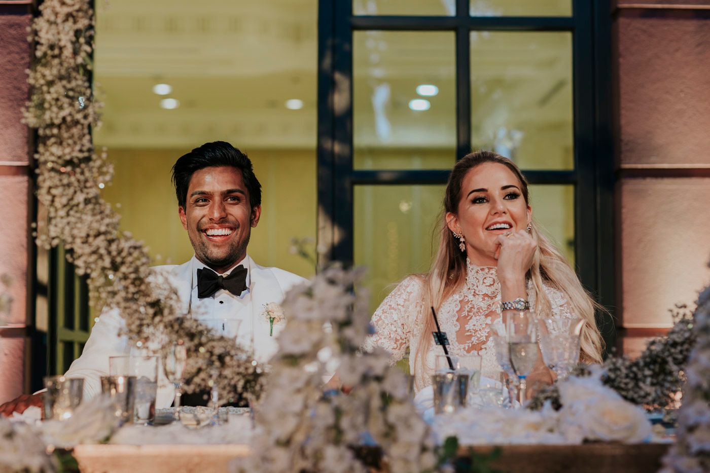 Two wedding guests at a decorated reception table appear behind lush floral arrangements and glowing candlelight.